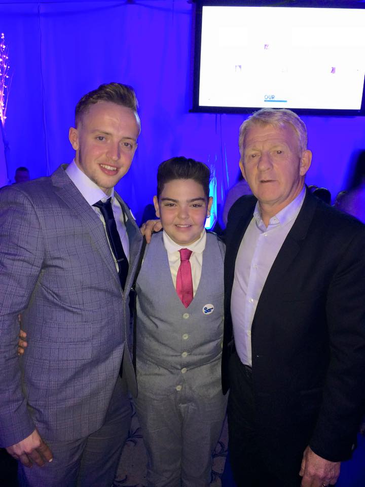 sam standing in a suit with 2 other men also dressed in suits